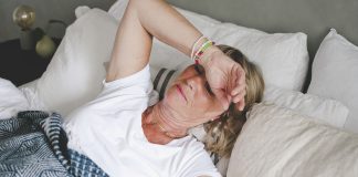 Mature white woman lies in bed with her arm over her face.