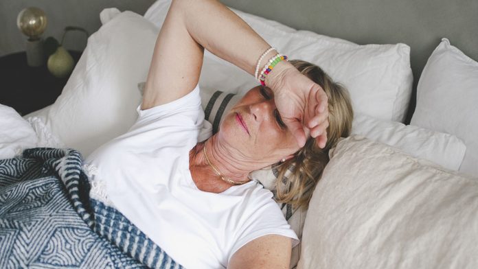 Mature white woman lies in bed with her arm over her face.